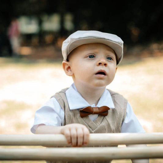 Baby boy wearing tan a flat cap, vest, and a leather bow tie standing behind a railing with a blurred background
