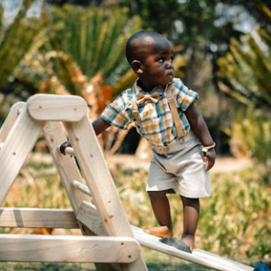 Baby boy wearing a check shirt with tan leather bow tie, suspenders and tan chino's playing on a wooden slide with a blurred natural background