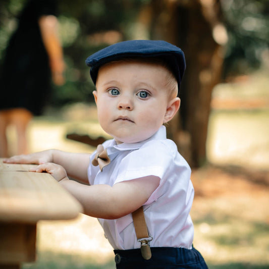 Baby boy wearing a navy flat cap and Tan leather suspenders in an outdoor setting