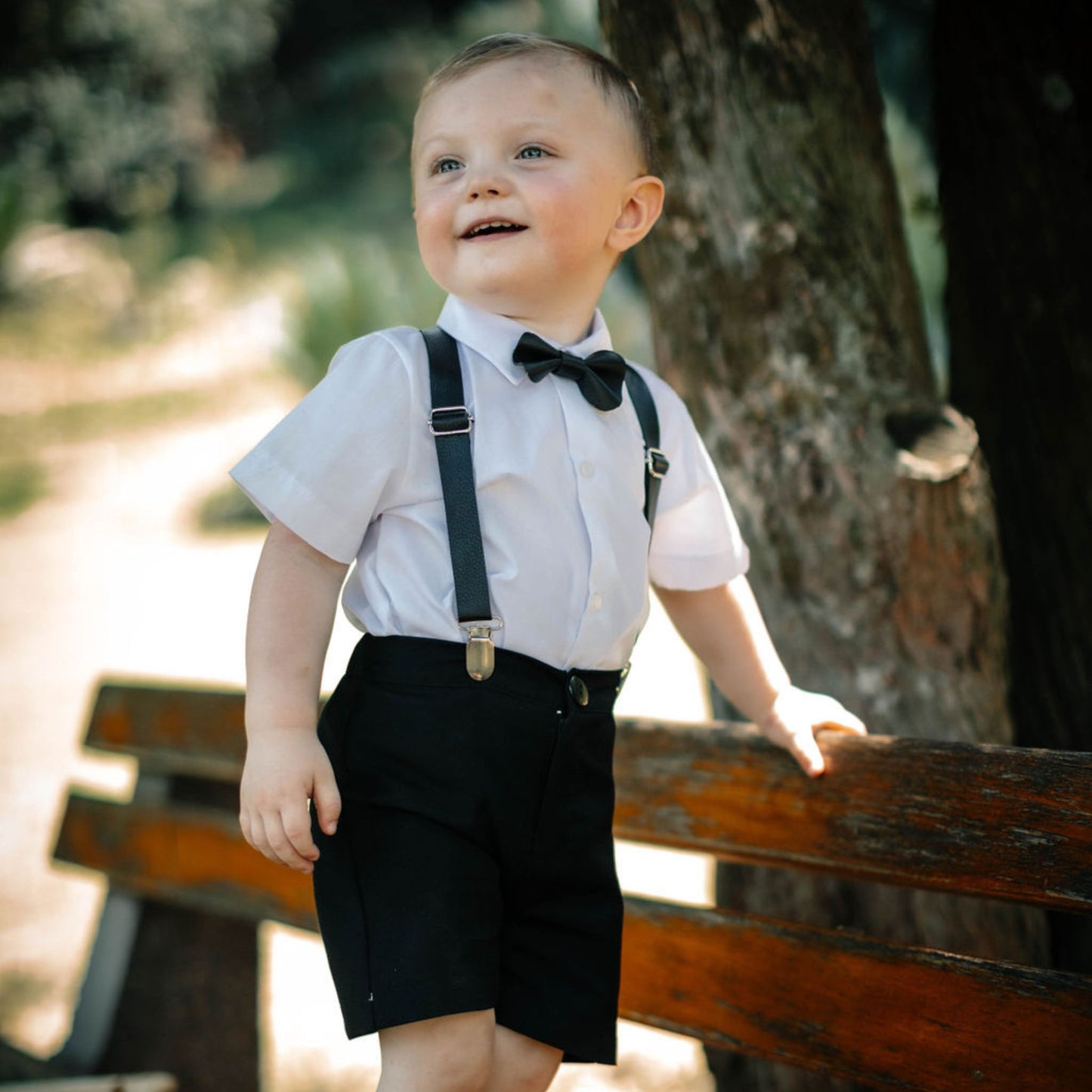 Baby in formal attire with black leather suspenders and bow tie standing outdoors on a bench