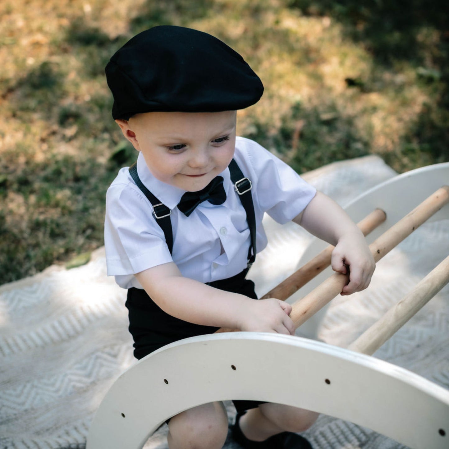 Baby wearing a white shirt with black leather bow tie and leather suspenders and a black flat cap playing on a wooden toy outdoors