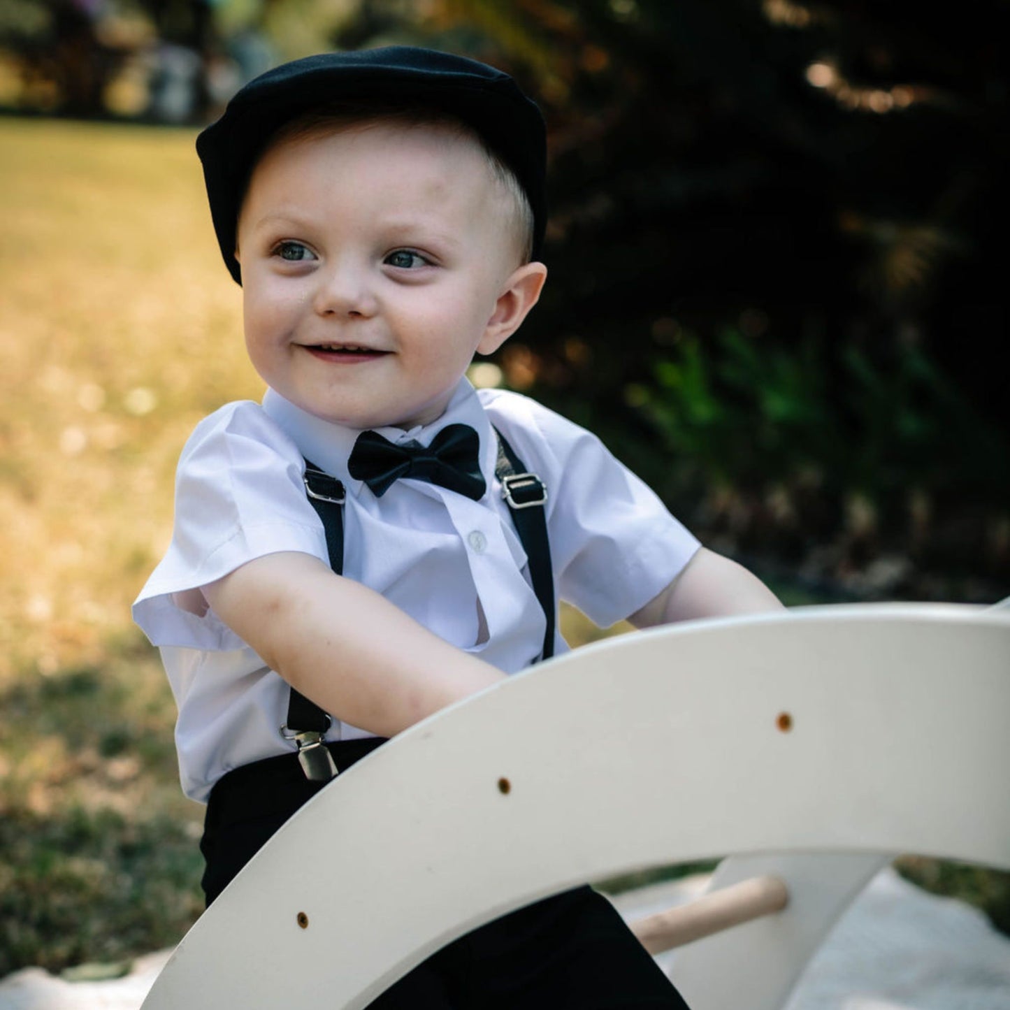 Baby wearing a white shirt, black leather bow tie, black leather suspenders and a black flat cap sitting on a wooden toy.