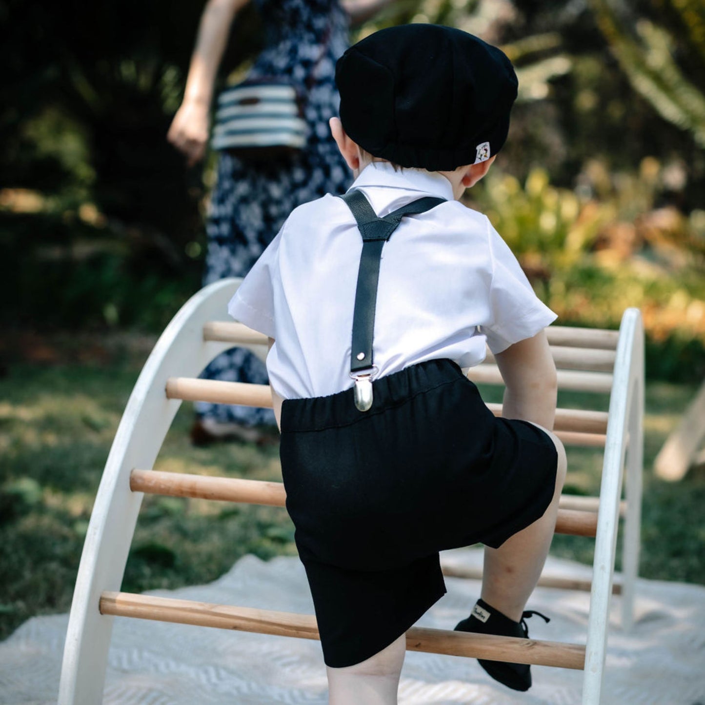 Baby from the back in a formal outfit with black leather suspenders, black leather shoes and a black flat cap climbing a ladder outdoors.