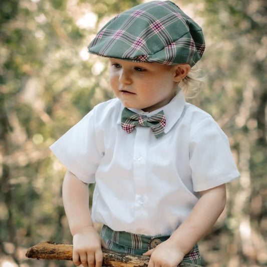 Baby wearing a plaid flat cap and bow tie, holding a stick with a forest background
