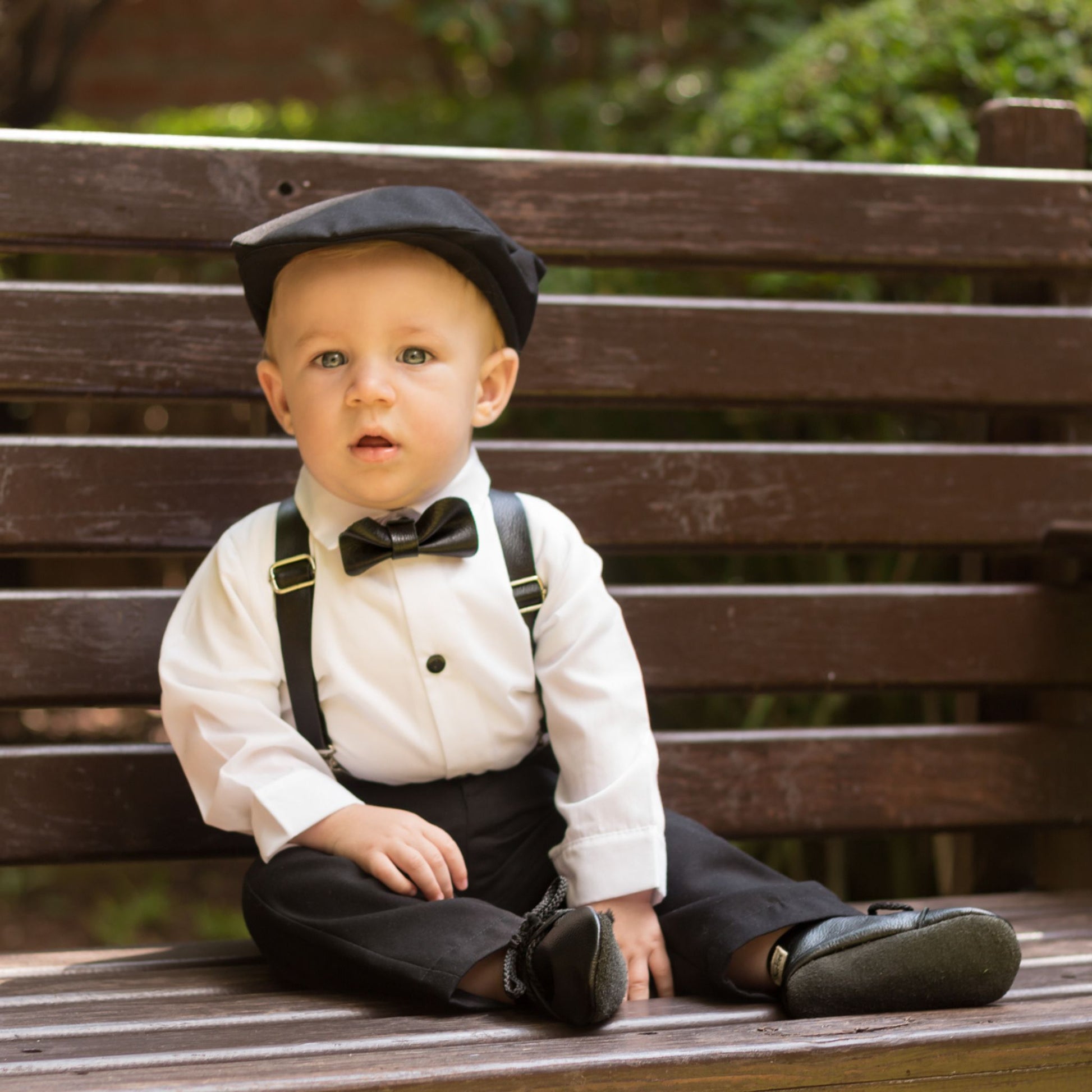 Baby boy wearing a black flat cap, leather bowtie, suspender and shoes  sitting on a wooden bench outdoors