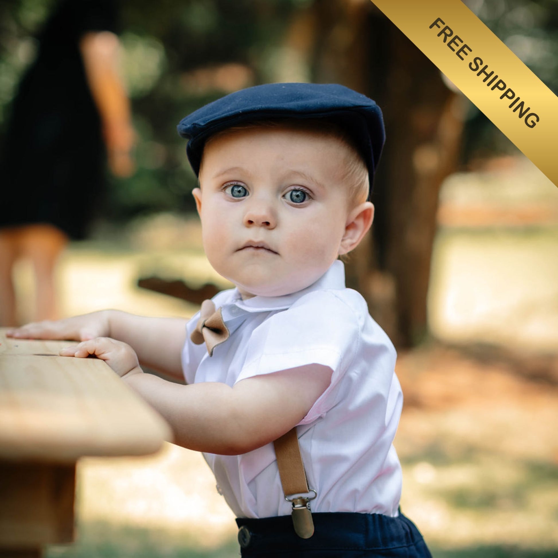 Baby boy wearing a flat cap and shirt with leather suspenders outdoors, with a free shipping banner overlay.
