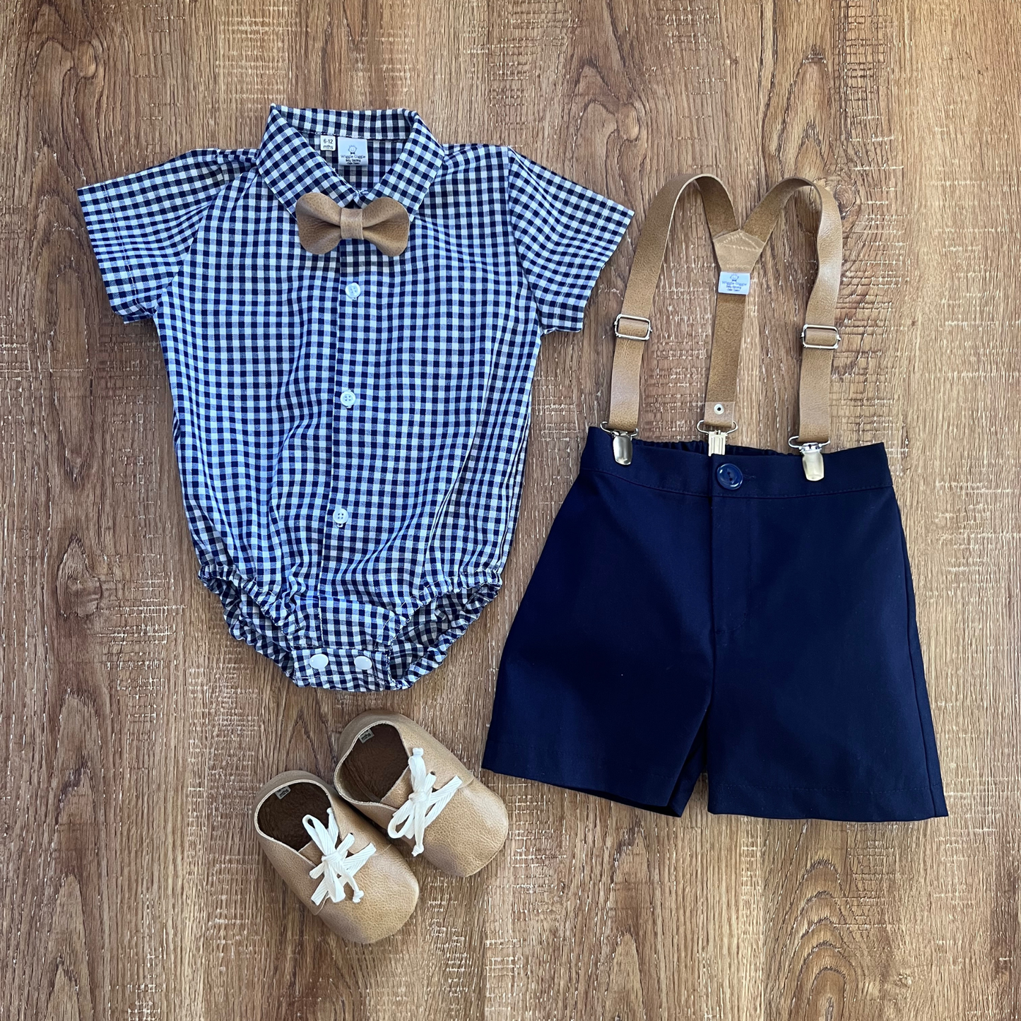 Baby boy outfit with checkered romper, navy shorts, suspenders, and shoes on a wooden surface