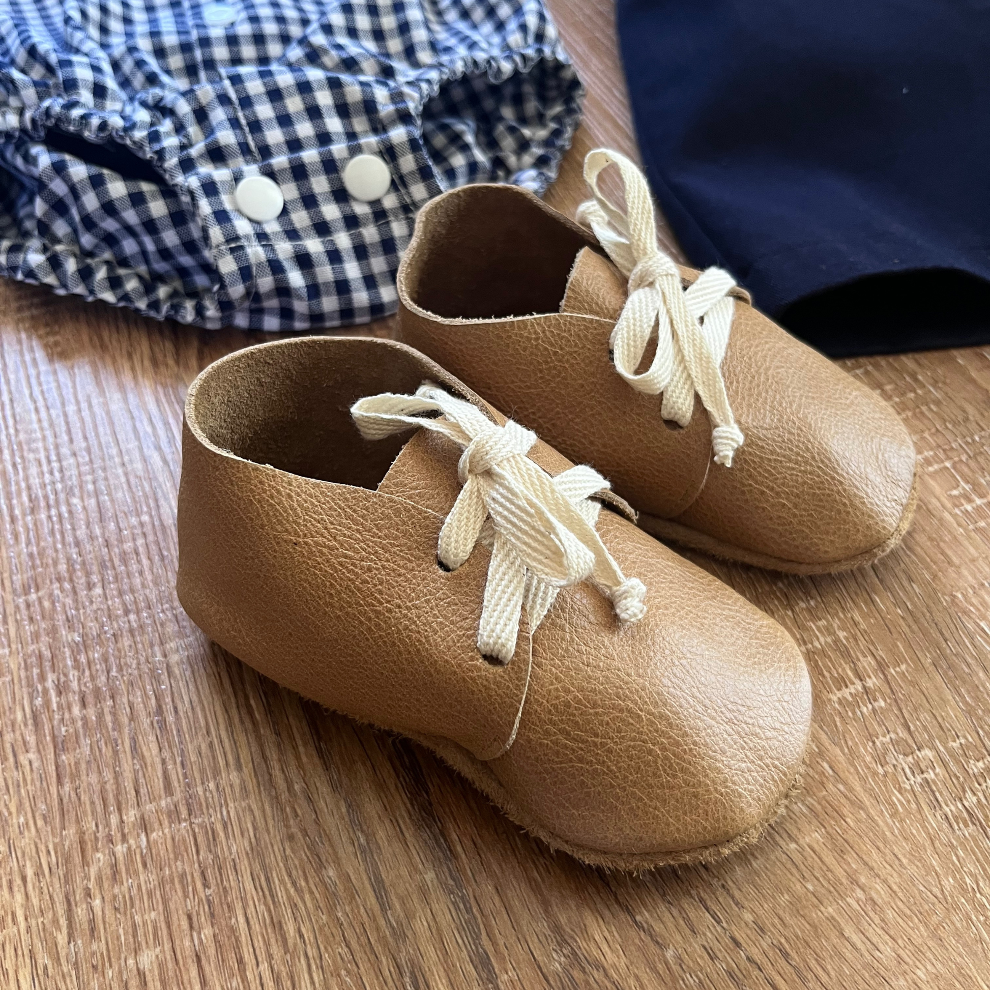 Pair of tan leather shoes with cotton laces on a wooden surface, next to blue checkered romper.
