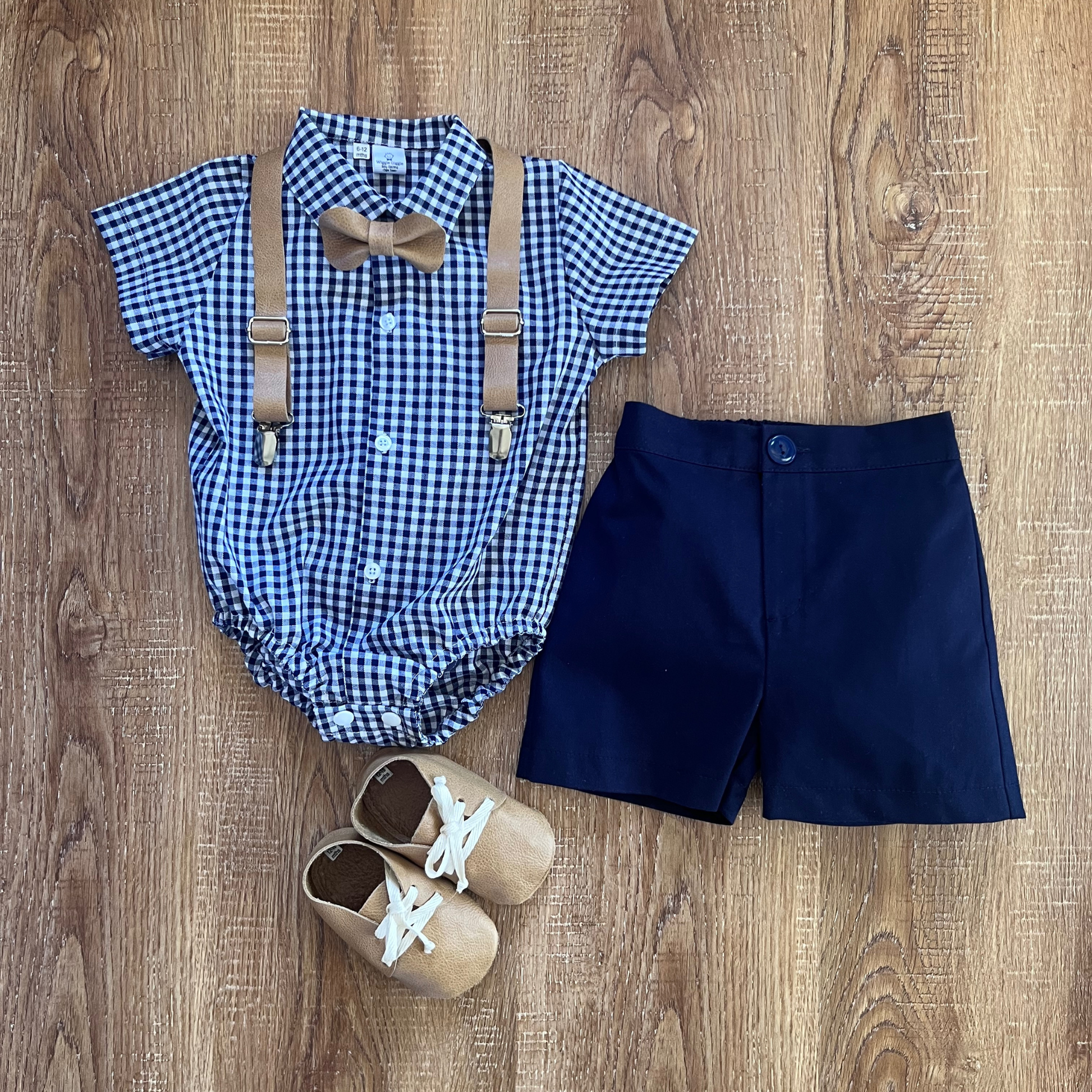 Blue checkered romper with suspenders, navy shorts, and baby shoes on a wooden surface