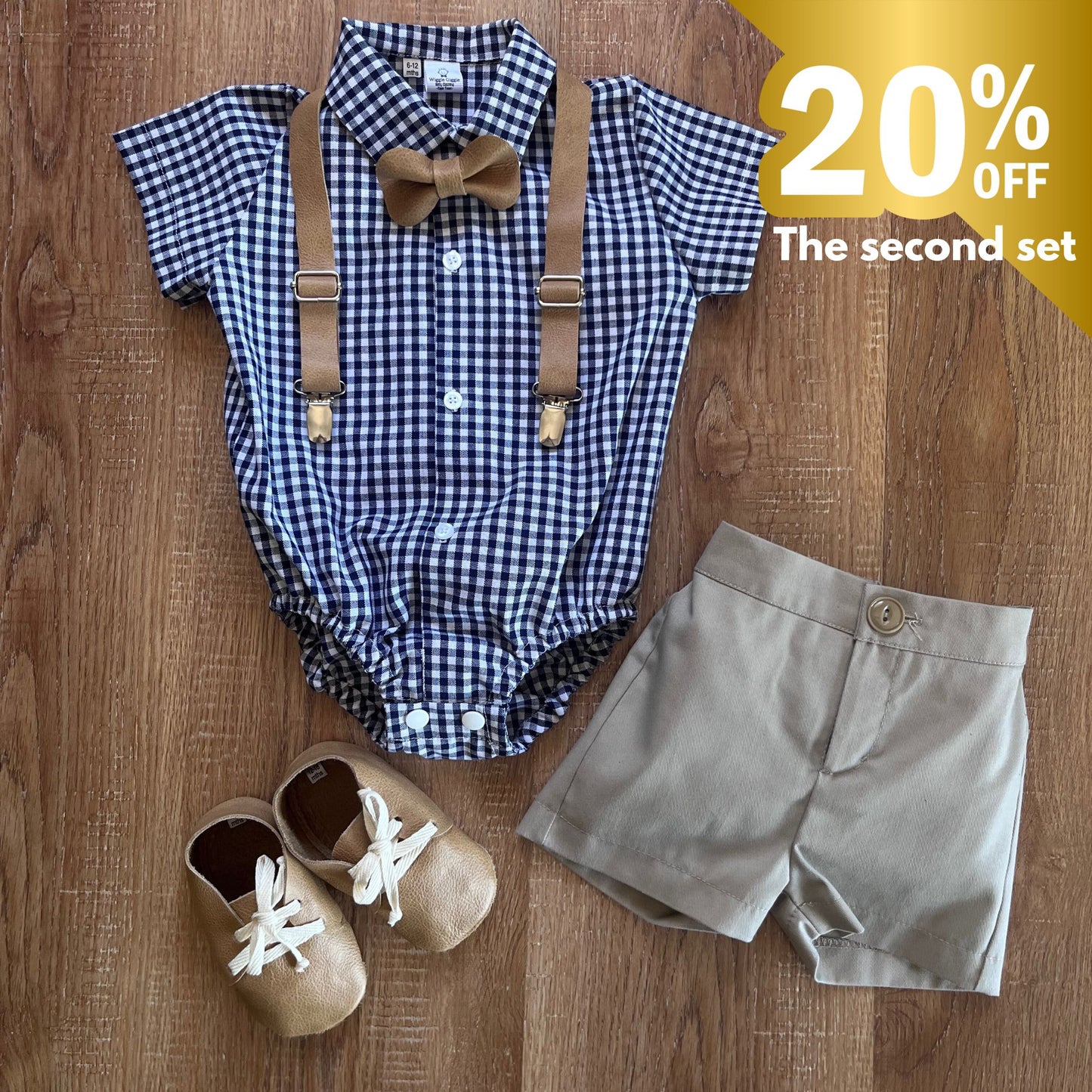 Baby boy outfit with checkered romper, suspenders, shorts, and shoes on a wooden surface with a discount label.
