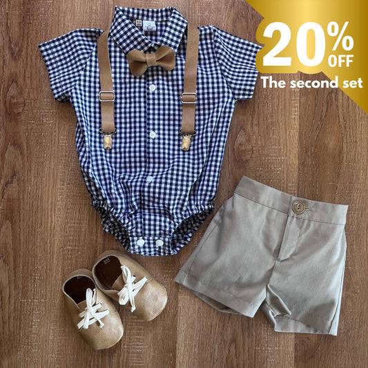 Baby boy outfit with checkered romper, suspenders, shorts, and shoes on a wooden surface with a discount label.