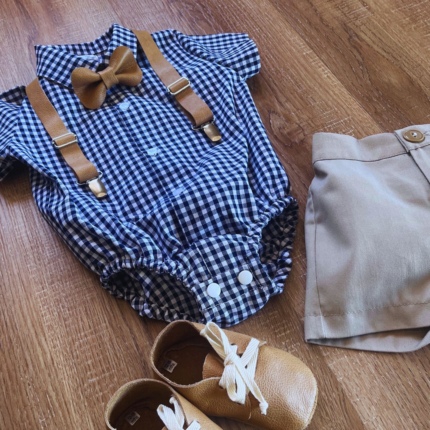 Baby boy outfit with blue checkered romper, tan leather shoes, beige pants, and leather bow tie on a wooden floor.
