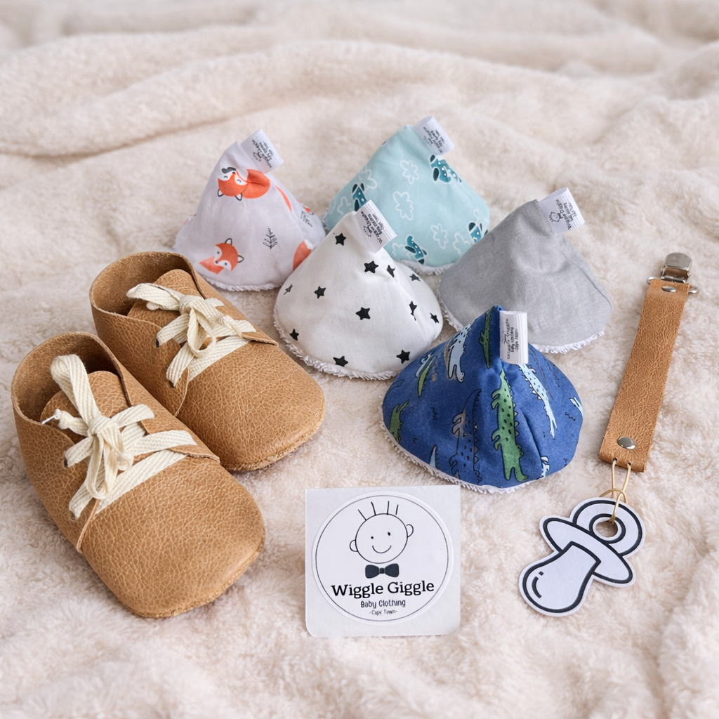 Baby leather shoes, leather pacifier clip and pee pee teepee's as a gift bundle with 'Wiggle Giggle' branding on a soft surface