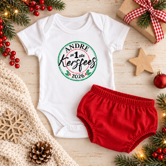 White onesie with a stamp design on the chest Andre se eerste Kersfees with a red diaper cover on a wood surface with Christmas decorations around it