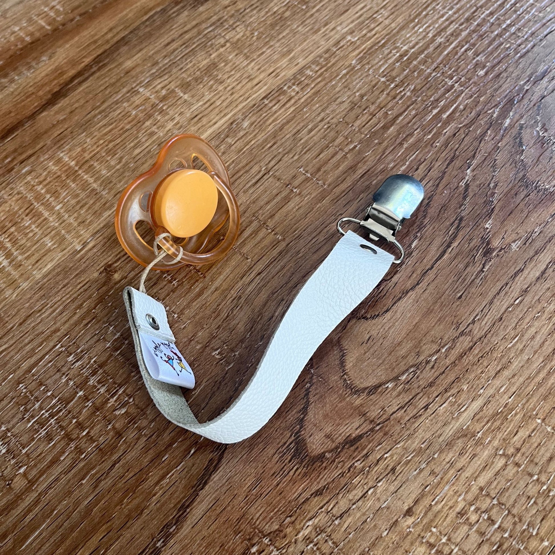 Orange pacifier with a white leather pacifier strap on a wooden surface