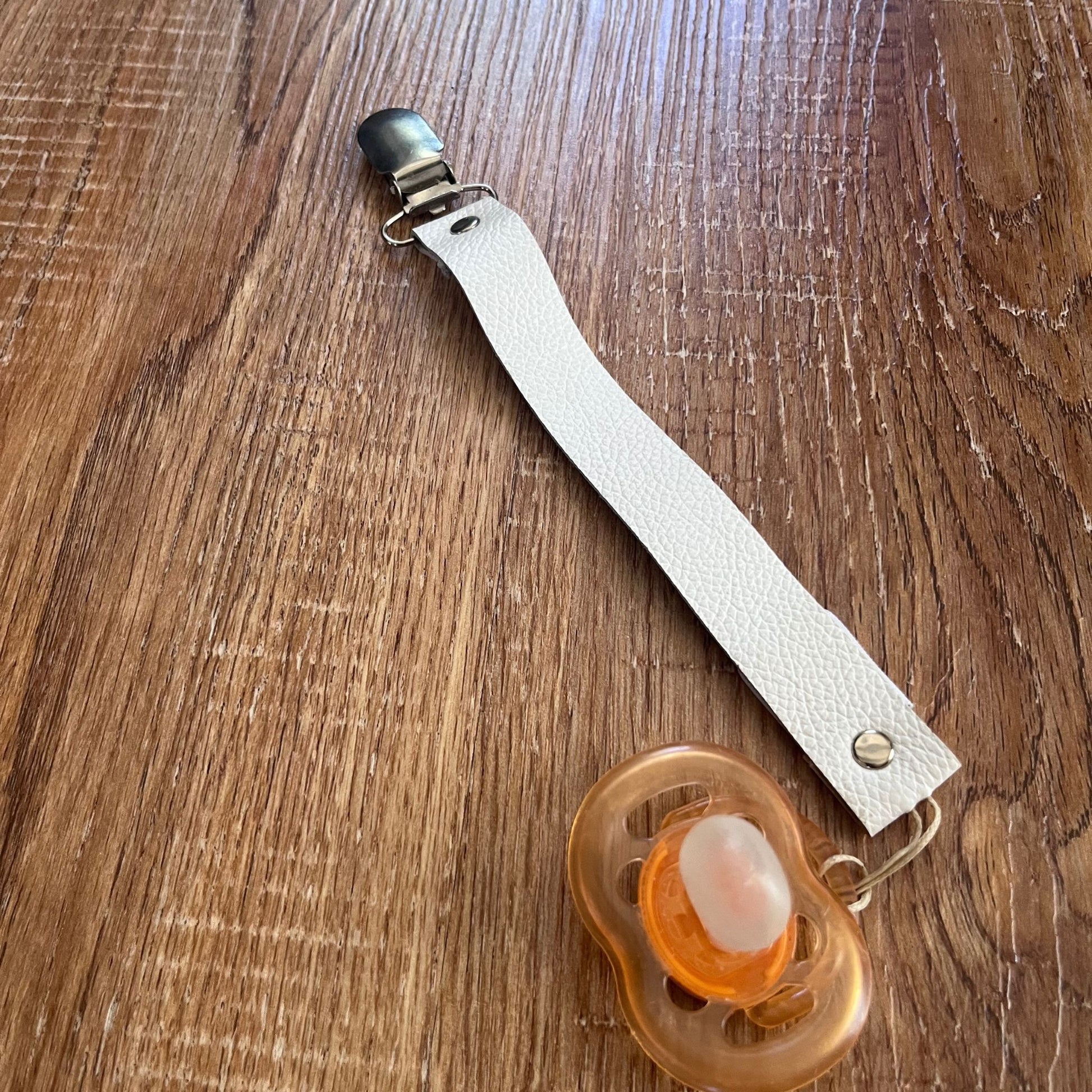 White leather pacifier strap with clip and orange pacifier on a wooden surface